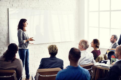 Eine weibliche Lehrkraft steht vor einem Whiteboard und redet. Vor ihr sitzen in einem Halbkreis Frauen und Männer verschiedenen Alters und hören ihr zu.