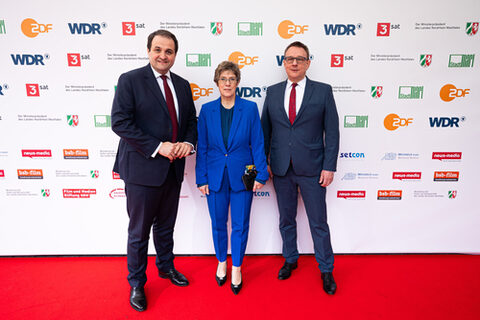 Drei festlich gekleidete Personen stehen auf dem roten Teppich vor einer Logowand und schauen lächelnd in die Kamera. In der Mitte eine Frau in blauem Anzug, links und rechts von ihr Männer in dunkelblauen Anzügen.