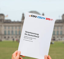 Im Vordergrund halten zwei Hände den Koalitionsvertrag "Verantwortung für Deutschland" von CDU, CSU und SPD hoch. Im Hintergrund ist verschwommen der Deutsche Bundestag in Berlin zu sehen.