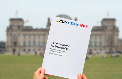 Im Vordergrund halten zwei Hände den Koalitionsvertrag "Verantwortung für Deutschland" von CDU, CSU und SPD hoch. Im Hintergrund ist verschwommen der Deutsche Bundestag in Berlin zu sehen.