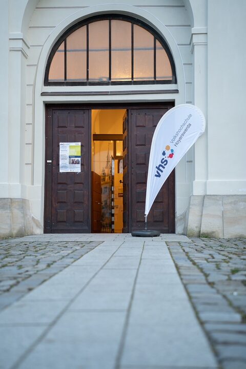 Eine geöffnete Holztür mit einem Fensterbogen darüber, Vor der Tür steht eine Fahne mit dem Logo der vhs Hoeyerswerda.