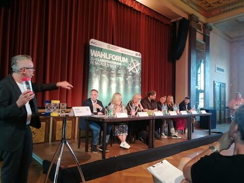 Auf der Bühne sitzen mehrere Personen an einem langen Tisch, vor dem sich Namensschilder befinden. Links im Bild steht ein Moderator mit einem Mikrofon und einer großen Sanduhr in der Hand. Im Hintergrund befindet sich ein Banner mit der Aufschrift "Wahlforum zur Landtagswahl in Sachsen 2024". Es ist ein Publikum anwesend, welches die Diskussion verfolgt.