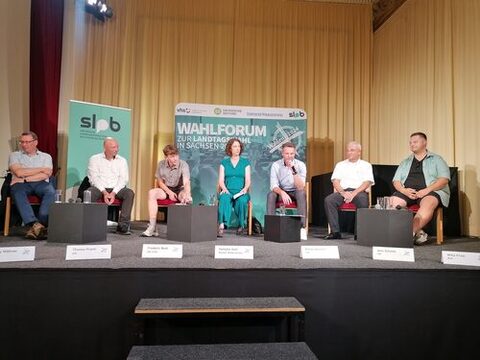 Sieben Personen sitzen auf einer Bühne, auf der sich im Hintergrund ein Banner mit der Beschriftung "Wahlforum zur Landtagswahl in Sachsen 2024" befindet. Von links nach rechts sind die Teilnehmer in einer Reihe auf Stühlen platziert. Vor jedem Teilnehmer steht ein Namensschild. Eine Person spricht in ein Mikrophon und die anderen hören zu.