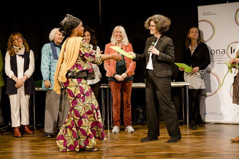 Eine Frau in mit schwarzem Kopfgewand, einer farbenfrohen Jacke und Rock nimmt ein Dokument von einer anderen Frau in einem schwarzen Anzug entgegen. Im Hintergrund stehen weitere Frauen.