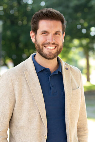 Ein Mann mit braunen Haaren und Bart mit beigefarbenem Sakko und dunkelblauem Poloshirt lächelt in die Kamera. Im Hintergrund sieht man verschwommen Bäume.