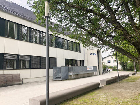 Ein weißes vhs-Gebäude hinter einem großen Baum und einer Wiese