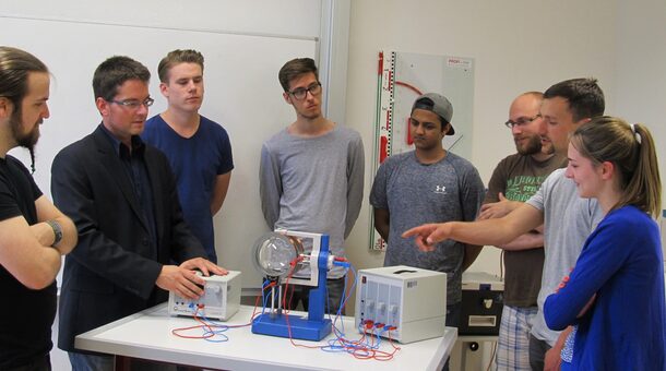 Eine Gruppe von jungen Männern und einer jungen Frau stehen um einen Tisch herum, auf dem elektrische Geräte aufgebaut sind.