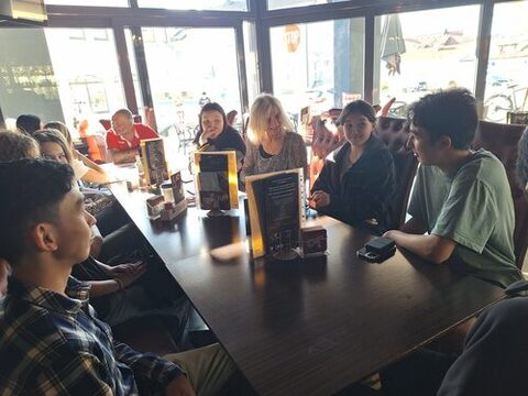 Eine Gruppe von männlichen und weiblichen Jugendlichen sitzt in einem Lokal. In ihrer Mitte eine ältere Frau mit blonden Haaren, die sich einem Jugendlichen zuwendet.