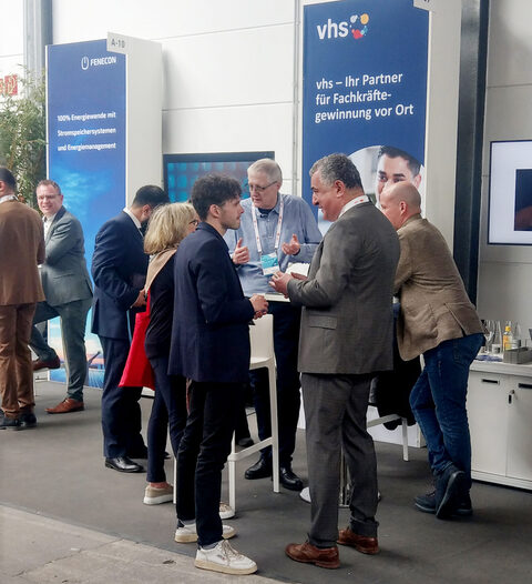 Männer und Frauen in Businessoutfits stehen an einem Messestand und unterhalten sich. Im Hintergrund ein blaues Banner mit dem bunten vhs-Logo und der Aufschrift "vhs - Ihr Partner für Fachkräftegewinnung vor Ort"
