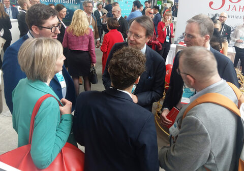 Eine Frau und mehrere Männer in Businessoutfits stehen inmitten einer Masse von Menschen in einer Halle und unterhalten sich.