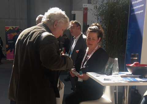 Eine Frau sitzt auf einem Hocker an einem Messestand und spricht mit einem älteren Mann.