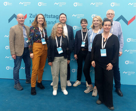 Vier Frauen und vier Männer in Business-Outfits stehen vor einer türkisfarbenen Fotowand und lächeln in die Kamera.