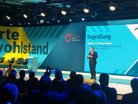 Ein älterer Mann im Anzug steht mit einem Mikro auf einer erleuchteten Bühne. Im Vordergrund Publikum, im Hintergrund grün-gelb gestaltete Wände und eine Projektion.