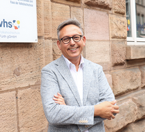 Ein Mann im grauen Jackett steht vor einer Gebäudefassade und lächelt in die Kamera. Hinter ihm sieht man ein Schild mit vhs-Logo und der Aufschrift "Volkshochschule Fürth gGmbh"