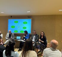 Blick auf ein Podium mit drei Frauen und zwei Männern, die vor einem Bildschirm mit Schaubild zu den vhs sitzen und mit dem Publikum sprechen.