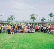 Eine große Gruppe von Menschen posiert aufgereiht auf einer grünen Wiese, im Hintergrund sieht man palmenartige Bäume und das Meer.