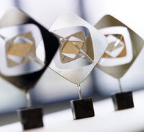 Drei Trophäen des Grimme-Preises nebeneinander aufgestellt in einem lichtdurchfluteten Saal