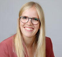 Eine junge Frau mit langen blonden Haaren, einer dunklen Brille und einem rötlichen Blazer lächelt in die Kamera.