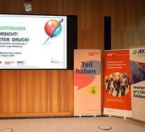 Ein Blick einer Leinwand auf die eine Folie mit dem Titel der Fachtagung "Vorsicht unter Druck!" projiziert ist. Daneben stehen drei Roll-Ups der Veranstalter Arbeit und Leben, Deutscher Volkshochschul-VErband und AKSB