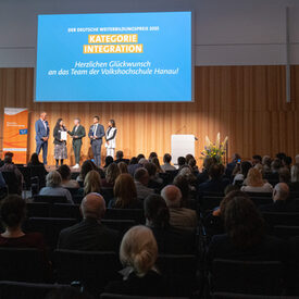 Blick über das Publikum hin zu einer beleuchteten Bühne, auf der drei Männer und zwei Frauen stehen.