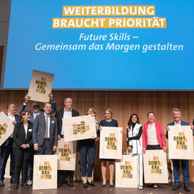Eine Gruppe von Männern und Frauen steht auf einer Bühne. Alle halten Schilder in den Händen, auf denen die Wörter "Bildung" und "Demokratie" stehen.