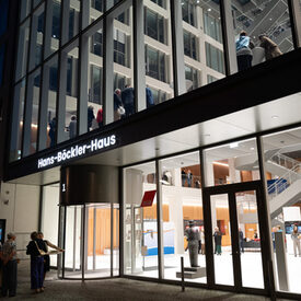 Außenansicht eines Gebäudes mit großer Fensterfront. Der Innenraum ist hell erleuchet.