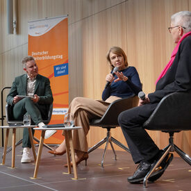 Ein Mann und zwei Frauen mit Mikrofon sitzen auf einer Bühne und diskutieren.