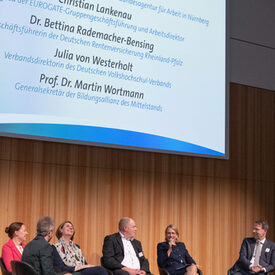 Drei Frauen und drei Männer sitzen auf einer Bühne. Eine Frau hält ein Mikrofon in der Hand.