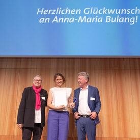 Zwei Frauen und ein Mann stehen auf einer Bühne. Die Frau in der Mitte hält einen Preis in der Hand.