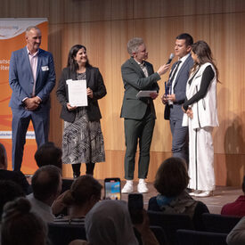 Zwei Frauen und drei Männer stehen auf einer Bühne. Eine Frau hält einen Preis in der Hand, der Mann in der Mitte hält dem Mann neben ihm ein Mikrofon hin.