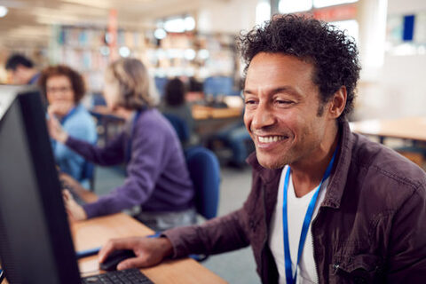 Ein erwachsener Mann sitzt bei einem Computerkurs an einem PC und lächelt.