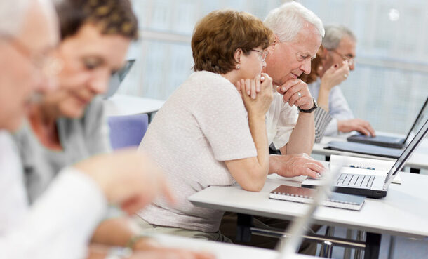 Ältere Frauen und Männer sitzen gemeinsam in einem Klassenraum an Laptops.
