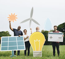 Vier Personen unterschiedlichen Alters stehen auf einer Wiese und halten große Papierschilder in der Form einer Glühbirne, einer Batterie, eines Windrades und einer Solarzelle in die Luft.