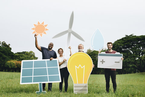 Vier Personen unterschiedlichen Alters stehen auf einer Wiese und halten große Papierschilder in der Form einer Glühbirne, einer Batterie, eines Windrades und einer Solarzelle in die Luft.
