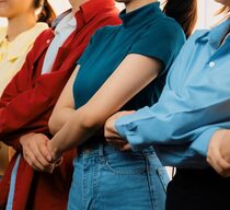 Personen unterschiedlichen Geschlechts in farbenfrohen Outfits stehen in einer Reihe zusammen mit überkreuzten Armen und gehaltenen Händen