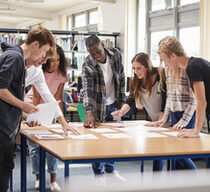 Eine Gruppe Jugendlicher steht an einem großen Tisch in einer Bibliothek und arbeitet gemeinsam an einer Idee.