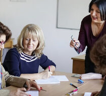 Weibliche Lehrkraft spricht mit erwachsenen Schülerinnen, die am Schreibtisch im Klassenzimmer sitzen.