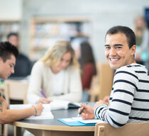 Studierender sitzt am Tisch und lächelt in Kamera