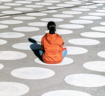 Symbolbild für Einsamkeit: Ein Mensch in einem orangefarbenen Hoodie sitzt auf einem großen Platz, auf dem viele weiße Punkte gedruckt sind