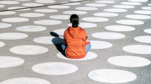 Symbolbild für Einsamkeit: Ein Mensch in einem orangefarbenen Hoodie sitzt auf einem großen Platz, auf dem viele weiße Punkte gedruckt sind