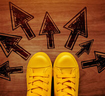 Symbolbild für Zukunftsvisionen: Gelbe Schnürschuhe auf einem Holzboden - um die Schuhe herum zeigen schwarze Pfeile in unterschiedliche Richtungen