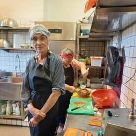 Zwei Frauen mit Tüchern um den Kopf in einer großen Küche. Die Frau im Vordergrund lächelt in die Kamera, die Frau im Hintergrund bereitet Essen zu.
