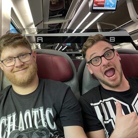 2 junge Männer sitzen in einem Zug und lachen in die Kamera