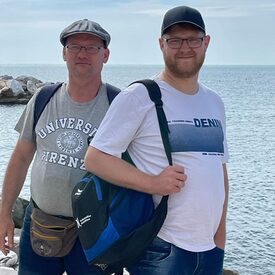 2 Männer posieren für die Kamera. Im Hintergrund das Meer und blauer Himmel.