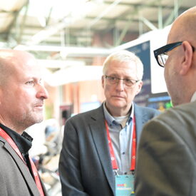 Drei Männer mittleren Alters im Gespräch in einer Messehalle