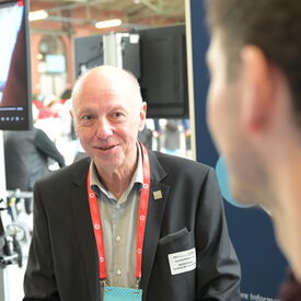 Älterer vhs-Vertreter vor Messestand mit Bildschirm nimmt Kontakt zu einem Messebesucher auf