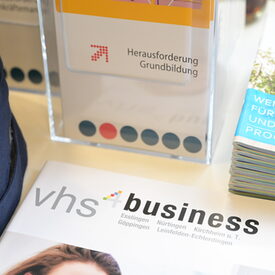 Detailaufnahme vom Messetisch. Zu sehen sind ein grauer Filzkorb, eine Broschüre "vhs4business", ein Dispenser für Flyer und ein Stapel Programmhefte
