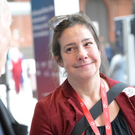 Eine vhs-Vertreterin mittleren Alters in weinrotem Cordsakko lächelt einen Messebesucher an