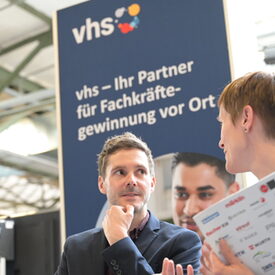 Ein DVV-Vertreter mittleren Alters schaut eine Messebesucherin mittleren Alters an, die ein Heft in der Hand hält und sich ihm gestikulierend zuwendet