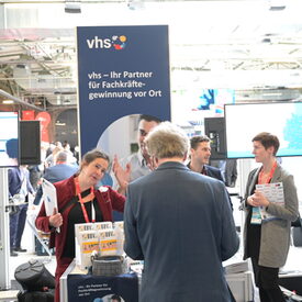 Messesituation mit Blick auf zwei Bildschirme und einem vhs-Messebanner in der Mitte. Davor ein Tisch mit Flyerdispensern. Hinter dem Tisch stehen drei Frauen und ein Mann, vor dem Tisch steht ein Messebesucher.